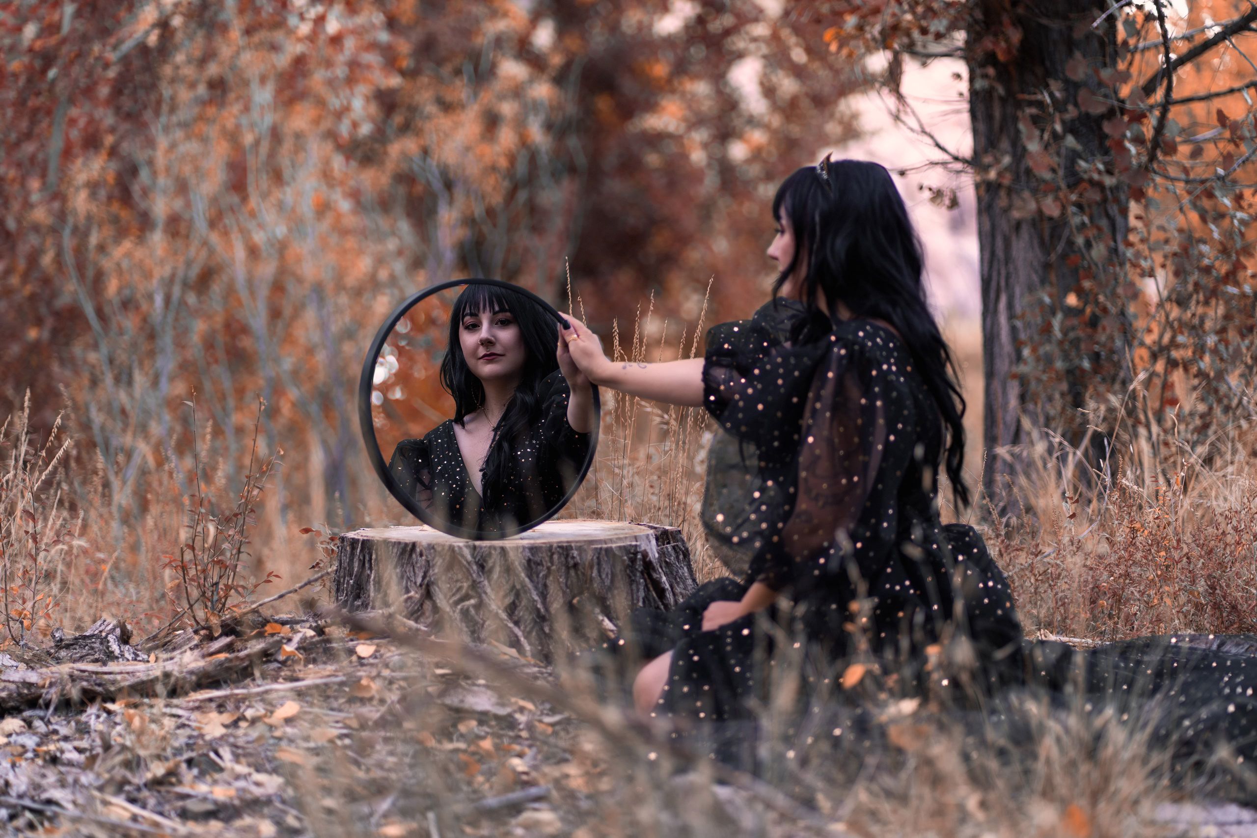 Woman in a celestial black gown looking into a round mirror on a tree stump in autumn woods